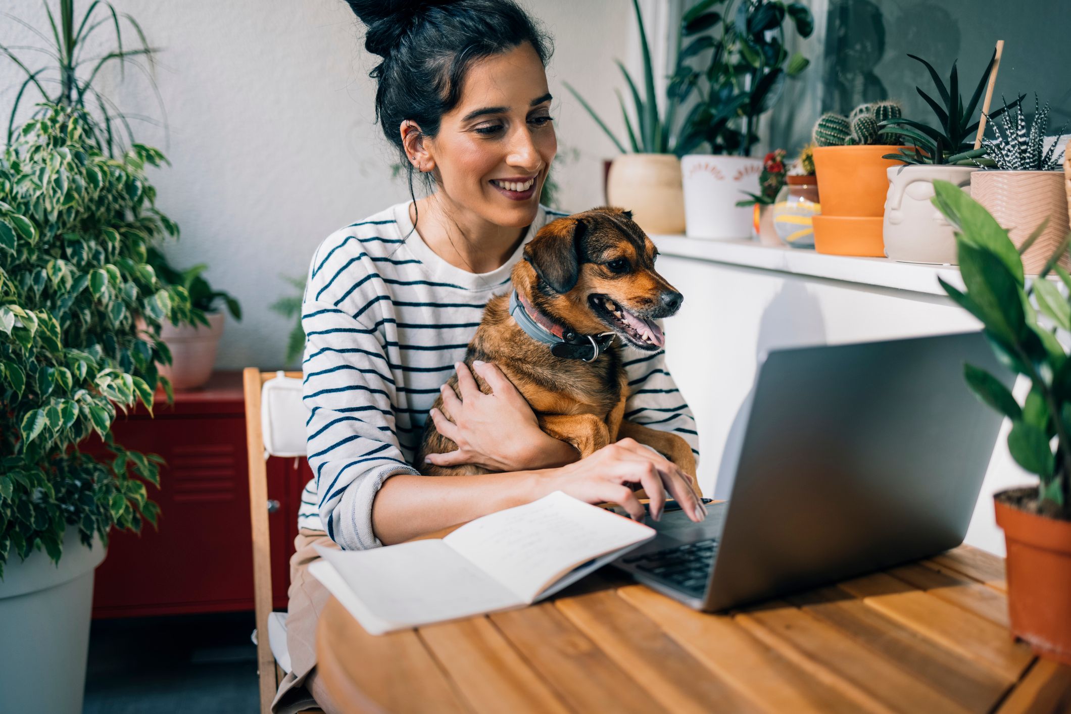 Woman holding her dog and smiling while typing on a laptop for a virtual vet appointment.