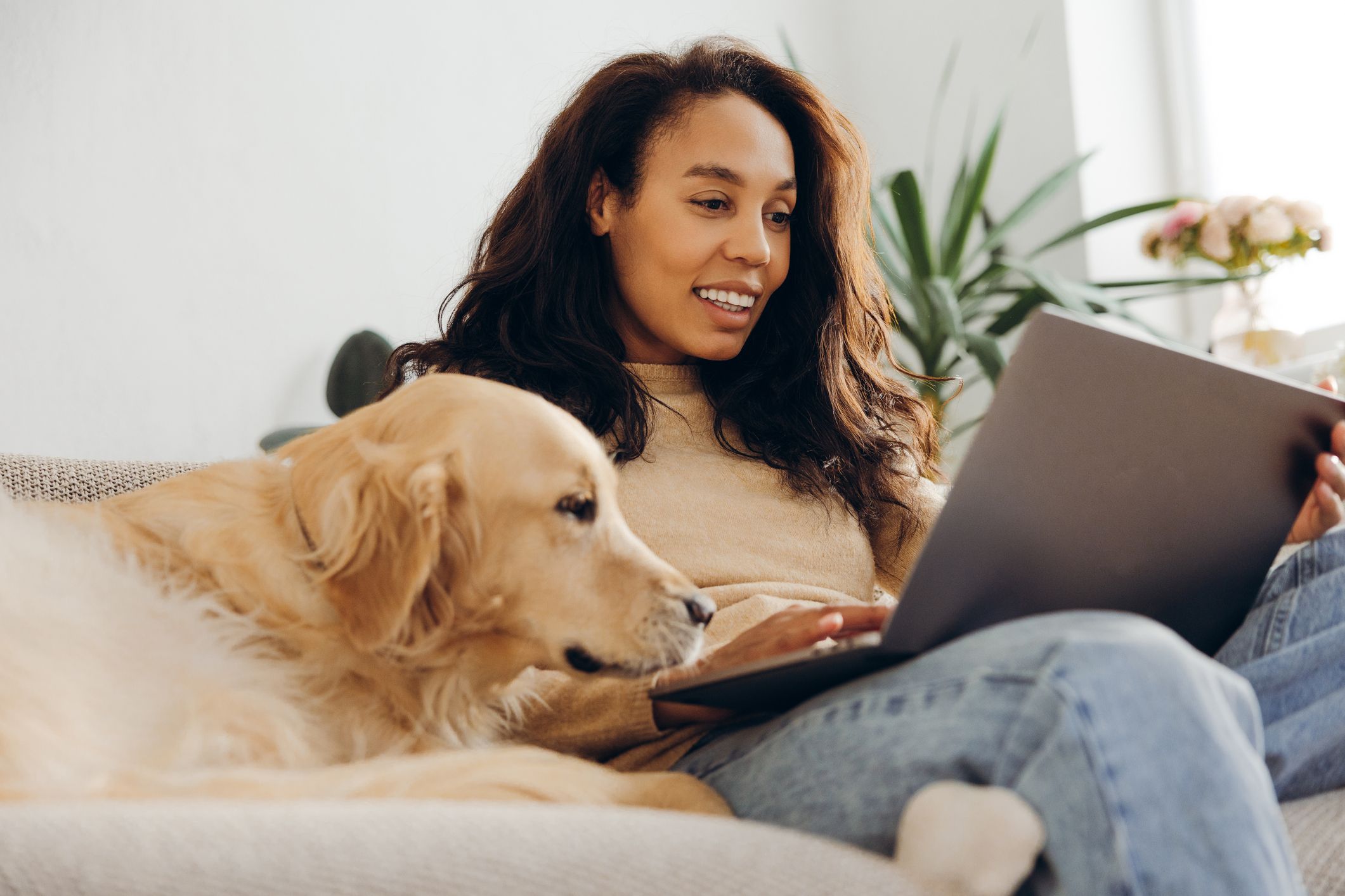 Woman on a couch with her golden retriever, smiling at her laptop.